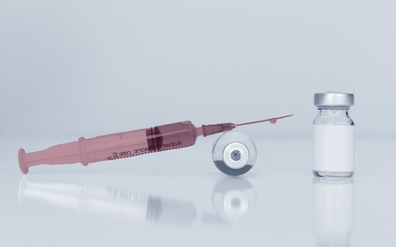 eine Spritze, an deren Nadel ein Tropfen herunterläuft, liegt auf einem liegenden Vial (Injektionsfläschchen)